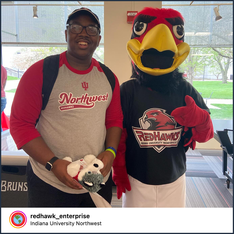 A man wearing a red-sleeved raglan T-shirt that features the IU Northwest logo smiles while standing beside IU Northwest mascot, Rufus the RedHawk.