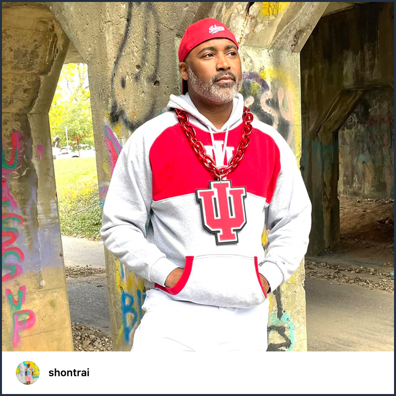 A man wears a red IU hat backwards, gray IU sweatshirt, and large chain necklace with a jumbo IU trident ornament while standing outside against a concrete pillar that’s tagged with multi-colored spray paint.